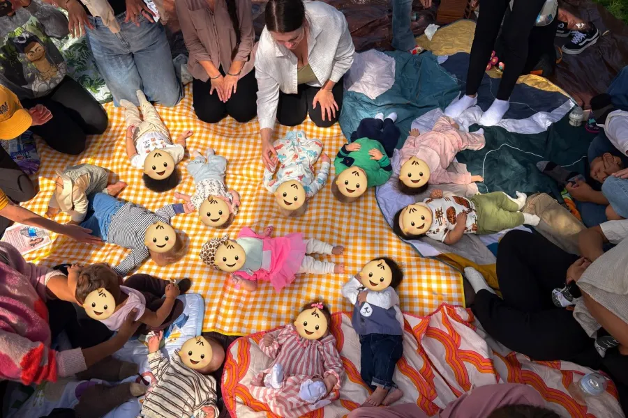 Babies forming a playful circle on a yellow picnic blanket, with their faces covered by emoji for privacy