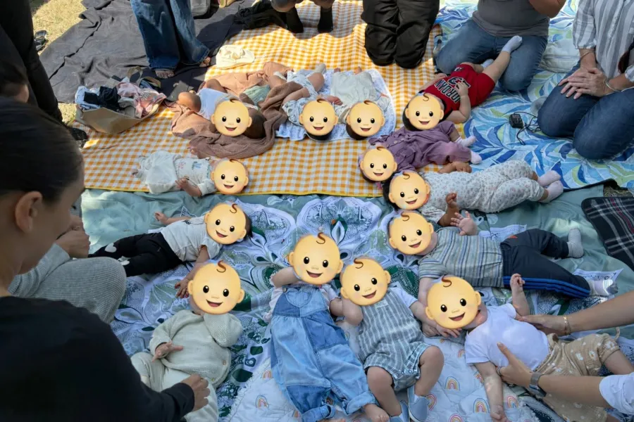 Babies lying together on picnic blankets while moms gather around them, with their faces covered by emoji for privacy